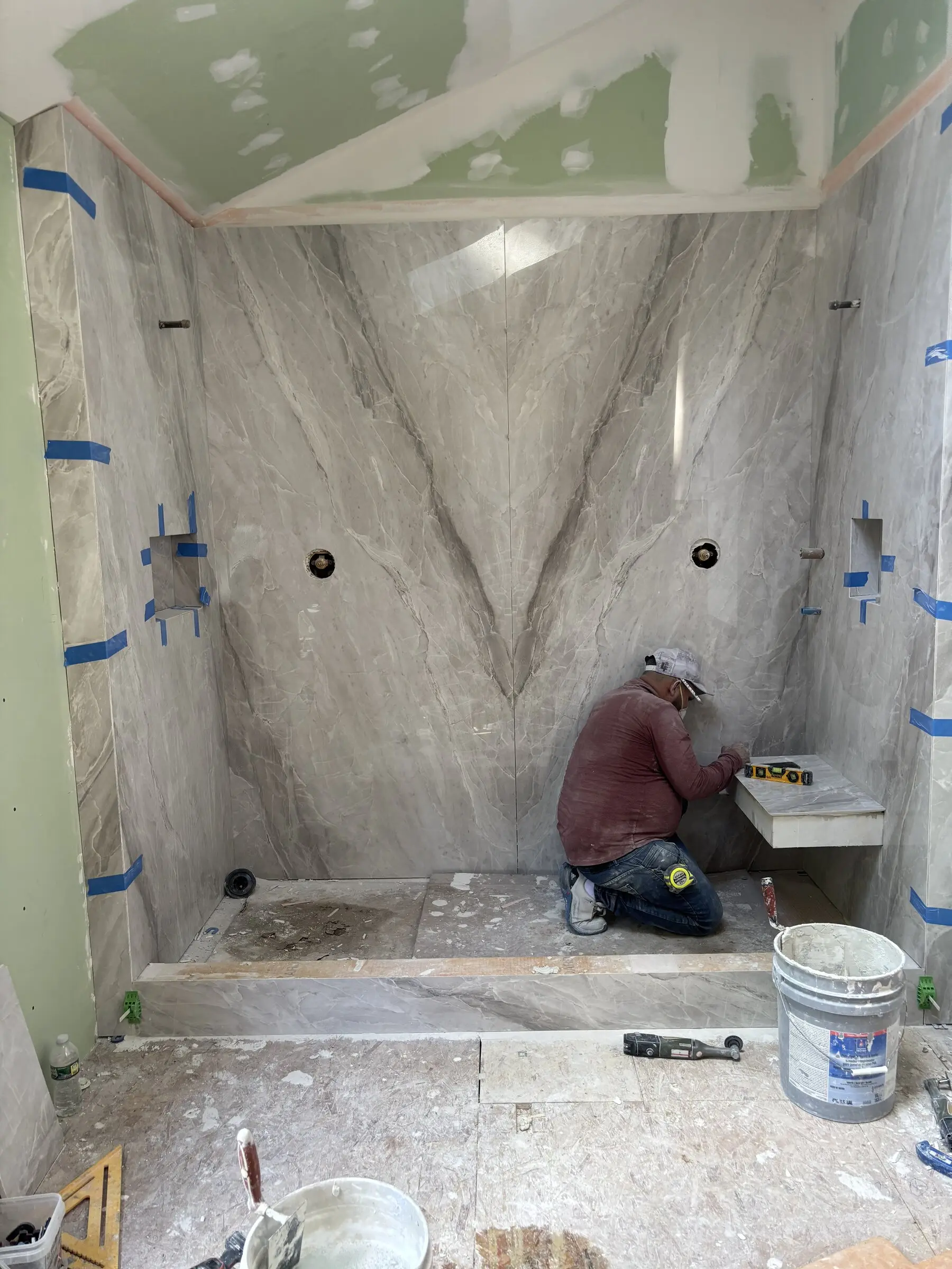 A Benchmark tile setter fitting the bench inside a book-matched marble shower, mid-build